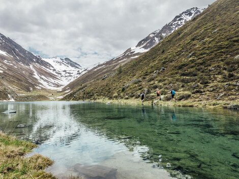 Ein ruhiger See in den Bergen, umgeben von schneebedeckten Gipfeln. Wanderer stehen am Ufer und genießen die Natur.