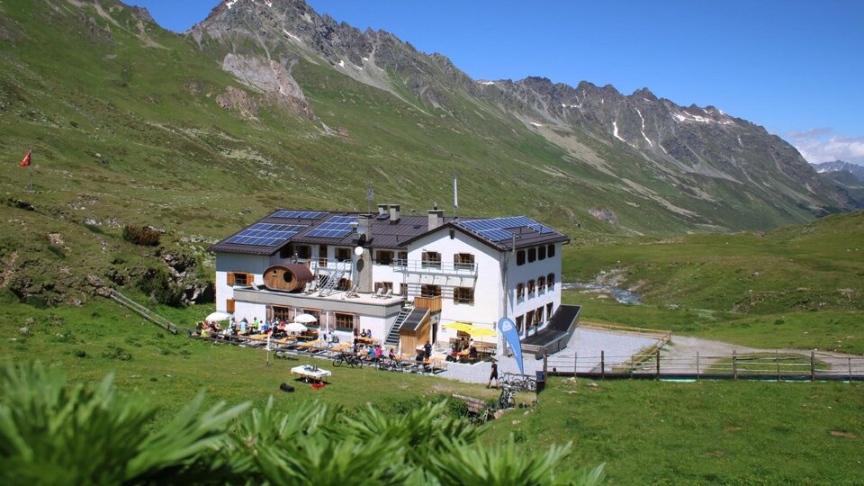 A charming mountain house in a green landscape. In the background, majestic mountains and a blue sky are visible.