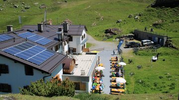 A rustic building with solar panels in a green mountain landscape. Outside, there are tables with umbrellas for guests.