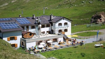 A mountain hut with terraces and guests sitting outdoors. Above the building are solar panels and the surroundings are green and hilly.