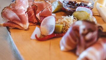 A beautiful wooden board with various types of sausage and cheese. Alongside are olives and a dip arranged in a small bowl.