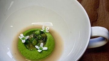 An elegant dish in a white bowl with a green cream and edible flowers. The background is made of wood, giving the image a rustic touch.