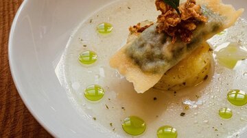An elegant dish served in a white bowl. It consists of ravioli with a filling, garnished with crispy onions and green droplets.