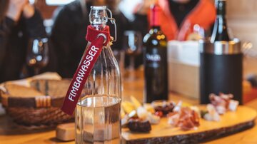 A glass bottle of water is on a table, surrounded by various wines and a selection of snacks. In the background, some people can be seen talking.