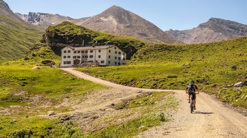 A cyclist is riding on a narrow gravel path in the mountains. In the background, there is a large, old building surrounded by green meadows and mountains.