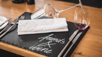 A stylishly set table with a wine glass and an overturned glass. A napkin and a black plate with the inscription "Friends & Wine" are placed on the table.