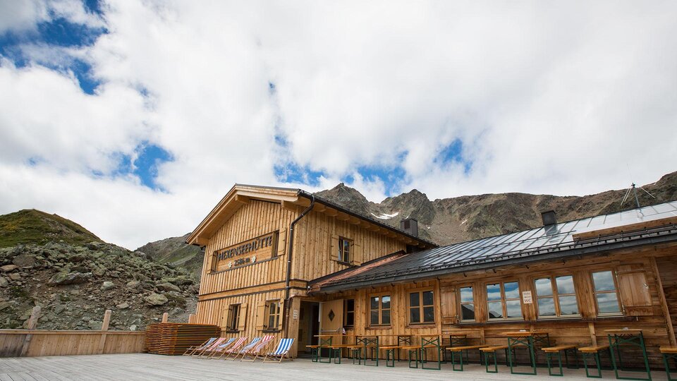 A rustic wooden house in the mountains with large windows. The sky is overcast and the surroundings are natural and green.