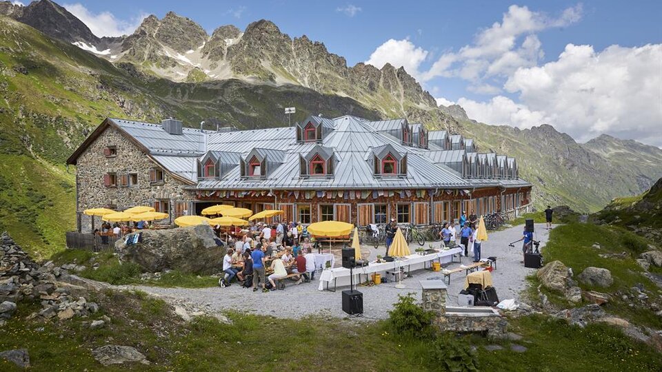 A cozy mountain cabin with outdoor seating and many guests. The surroundings are surrounded by impressive mountains and a clear blue sky.
