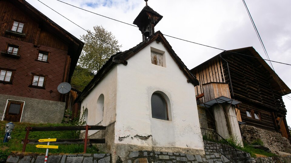 Eine kleine weiße Kapelle zwischen traditionellen Holzhäusern. Im Hintergrund sind Bäume und ein bewölkter Himmel zu sehen.