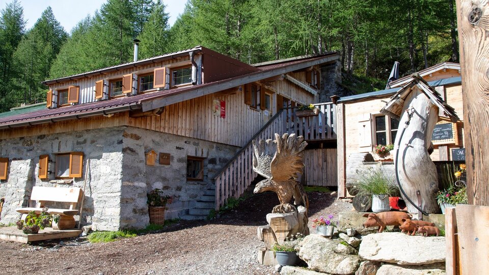 A cozy wooden house in the mountains, surrounded by green trees. In front of the house stands a decorative sculpture.