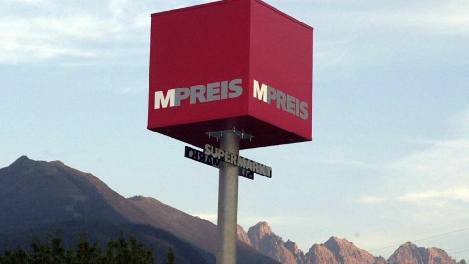 A red business sign with the inscription "MPREIS" in front of a mountain panorama. The sky is clear and the landscape is green.