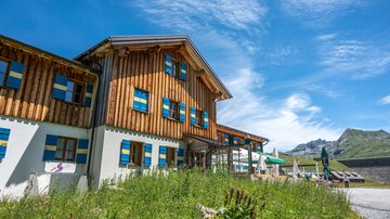 A cozy alpine wooden building in a natural setting. The sky is clear and the mountain landscape is visible in the background.