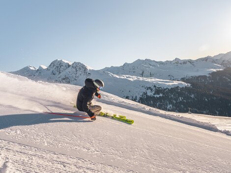 Ein Skifahrer fährt über eine verschneite Piste in den Bergen. Im Hintergrund sind beeindruckende Berglandschaften zu sehen.