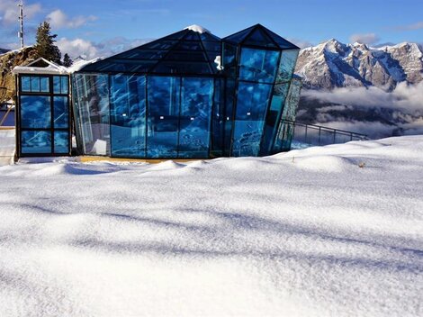 Ein modernes, gläsernes Gebäude steht inmitten einer schneebedeckten Landschaft. Im Hintergrund sind beeindruckende Berggipfel und ein klarer Himmel sichtbar.