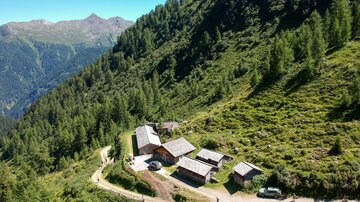 A picturesque mountain cabin surrounded by green meadows and trees. The path gently leads through the landscape into the mountains.