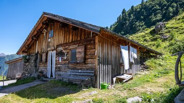 A traditional wooden cabin surrounded by green meadows and mountains. The sky is blue, and the sun is shining.