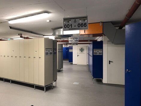 Ein moderner Umkleidebereich mit Spinden und geräumigen Gängen. Die Wände sind in Blau und Beige gehalten, und es gibt mehrere Nummernschilder.