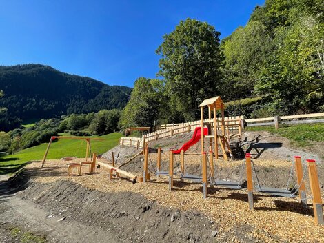 Ein Spielplatz in der Natur mit Holzelementen und einer Rutsche. Im Hintergrund sind grüne Hügel und Bäume zu sehen.