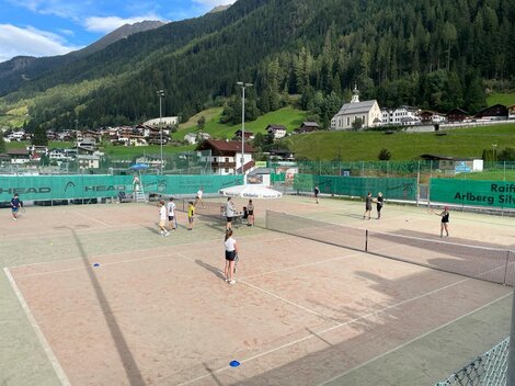 Ein Tennisplatz mit mehreren Spielern und einer bergigen Landschaft im Hintergrund. Die Umgebung ist grün mit typischer alpenländischer Architektur.
