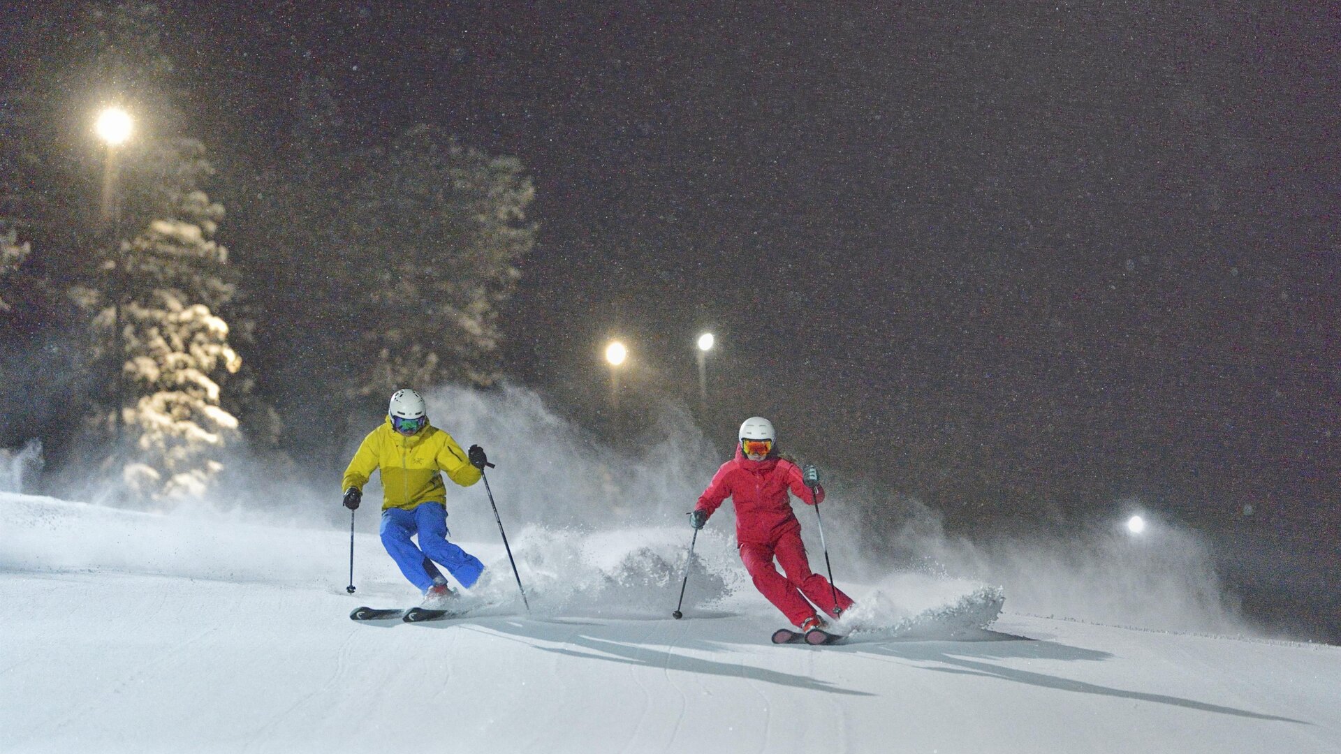 Two skiers are skiing at night on a snow-covered slope. The surroundings are illuminated, creating a winter atmosphere.