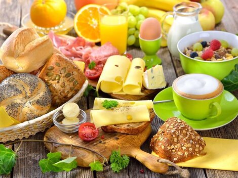 Ein reichhaltiges Frühstück mit verschiedenen Brotsorten, buntem Obst und Getränken. Dazu gibt es Käse und Joghurt in appetitlichen Schalen.