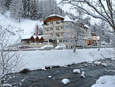 A picturesque winter scene with snow-covered trees and a clear river. In the background, a cozy hotel can be seen, surrounded by the snowy landscape.