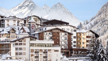 A picturesque winter village with snow-covered mountains in the background. Modern designed buildings and a clear, blue sky create a cozy atmosphere.