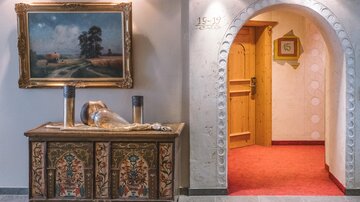 A stylish lobby with an ornate, painted cabinet and a painting on the wall. In the background, an arched passage leads to a room with red carpet flooring.